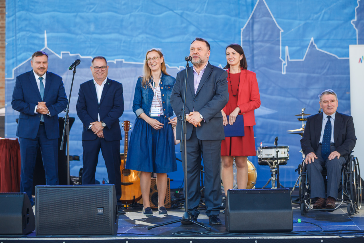 Scena plenerowa. Na pierwszym planie Zastępca Prezydenta Miasta Krakowa Andrzej Kulig. Stoi przed mikrofonem umieszczonym na statywie. Za nim stoją cztery osoby, a piąta jest na wózku inwalidzkim.